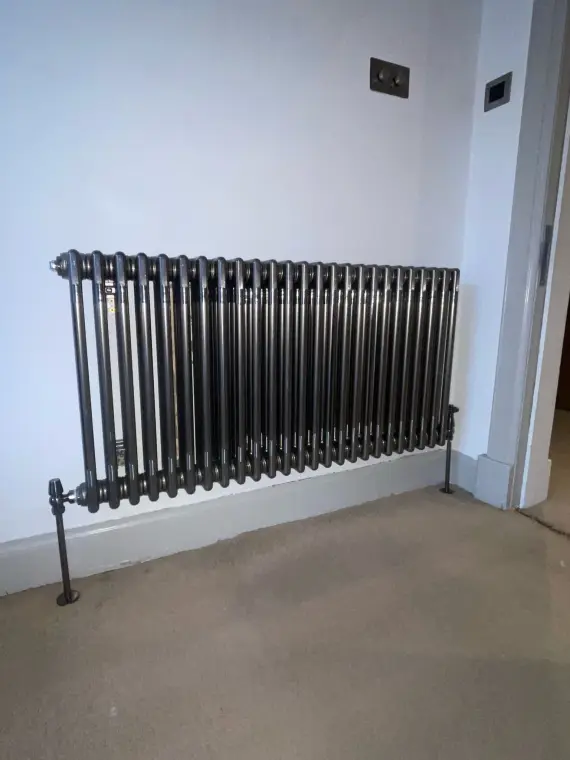 A gunmetal multi-column radiator fitted against a pale grey wall above a beige carpet.